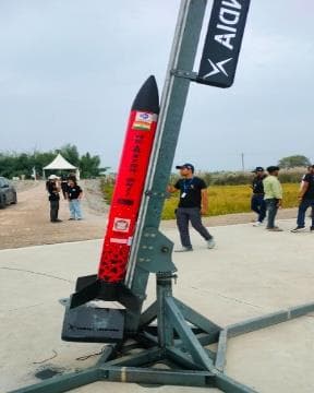 Red and black sounding rocket mounted on a steel launch rail at an outdoor launch site, with team members in the background.